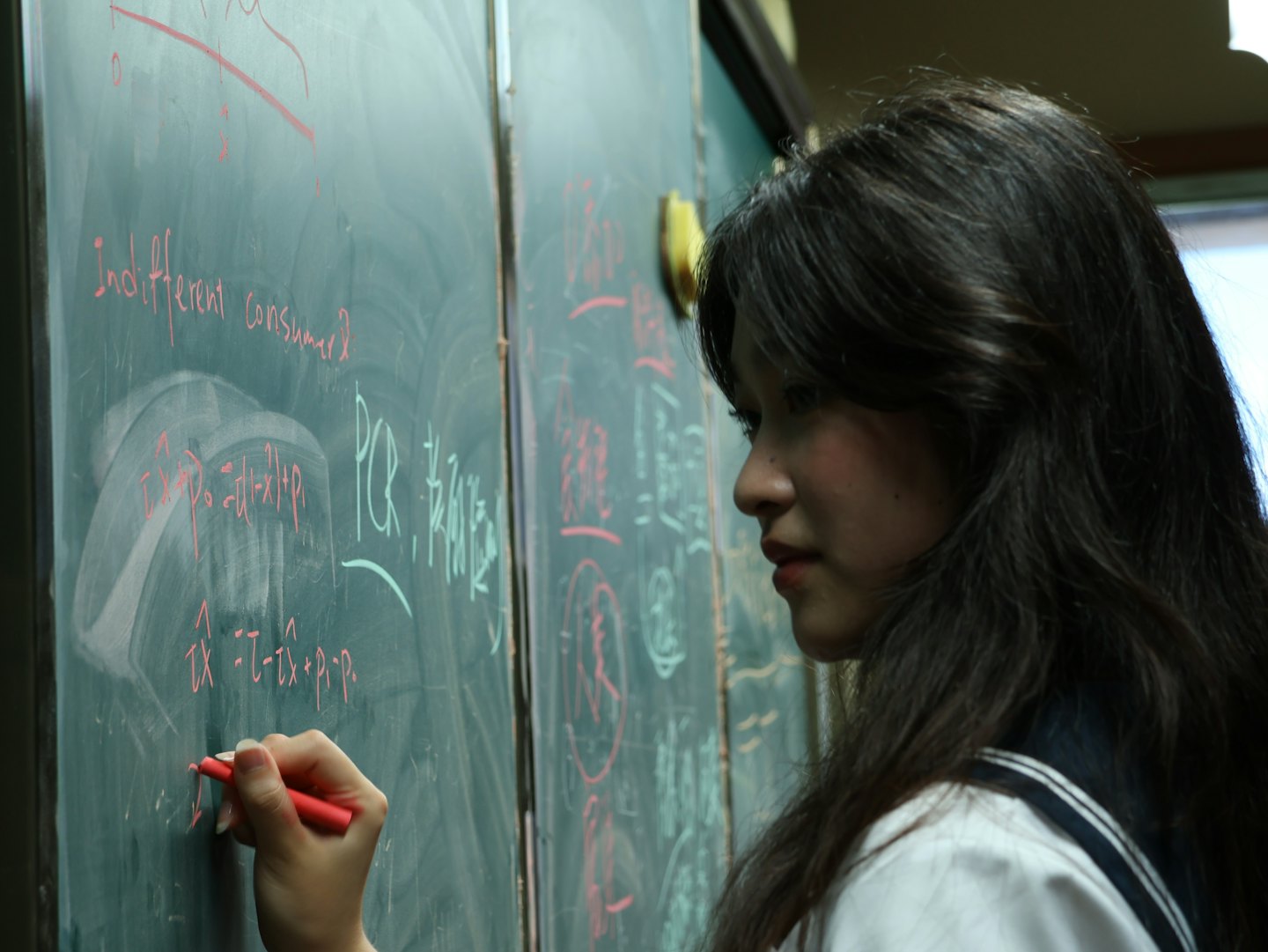 Woman in uniform writes equations on a blackboard with a red marker.