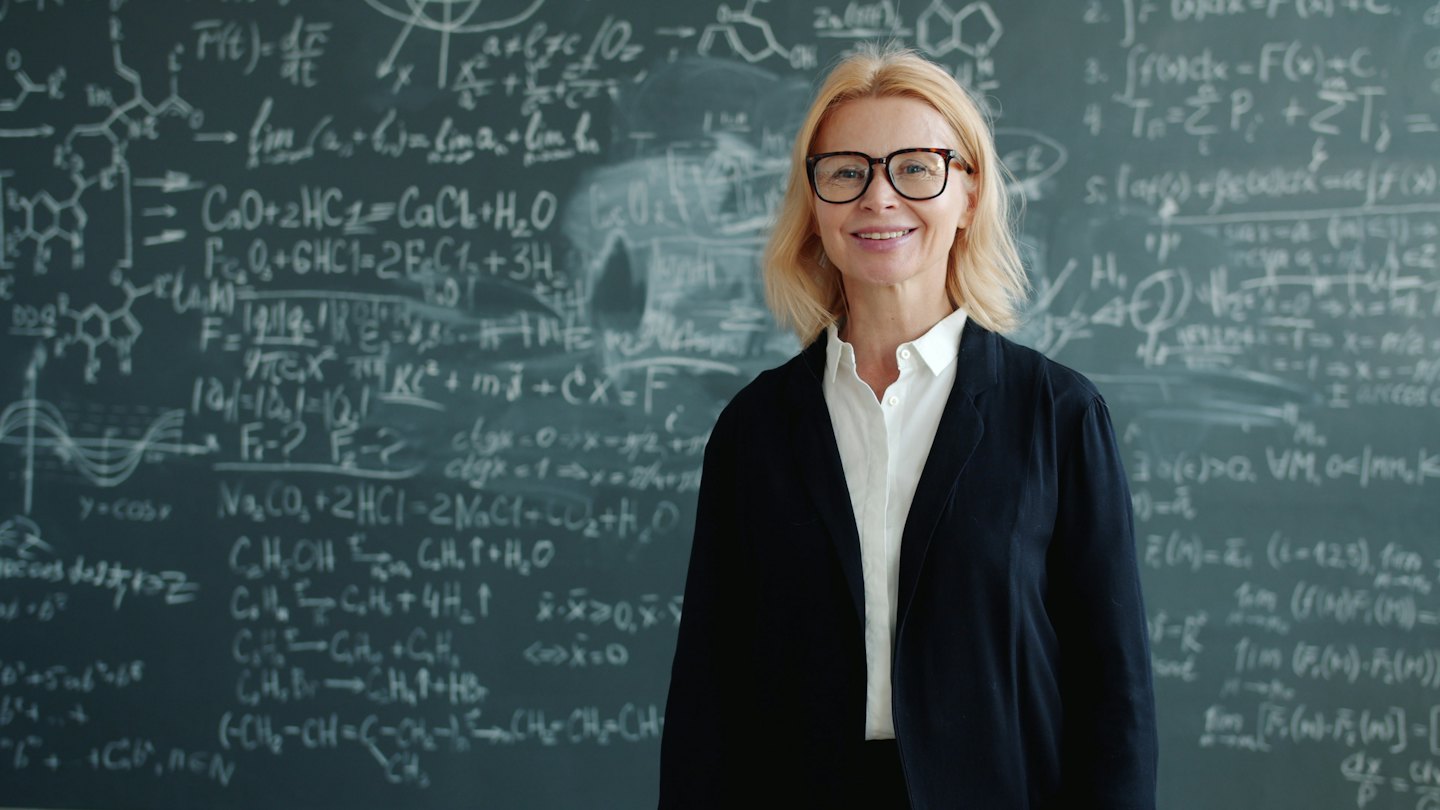 Woman smiles in front of chalkboard with science equations.