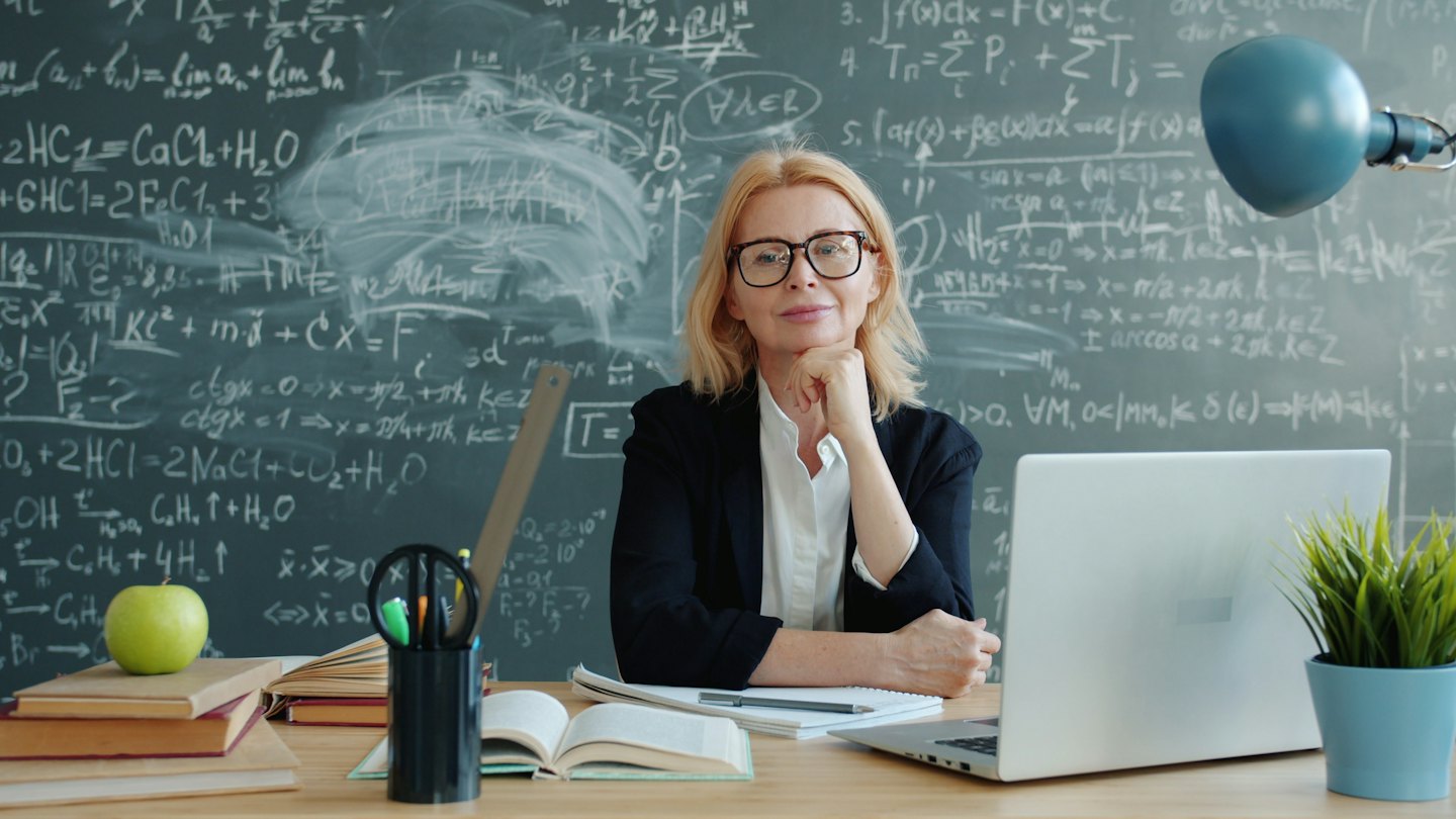 Woman at desk with laptop and chalkboard equations.